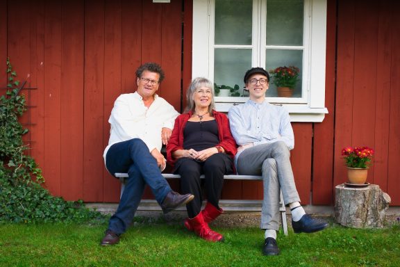 Tre personer sitter på en bänk framför ett rött hus med fönster och blommor.